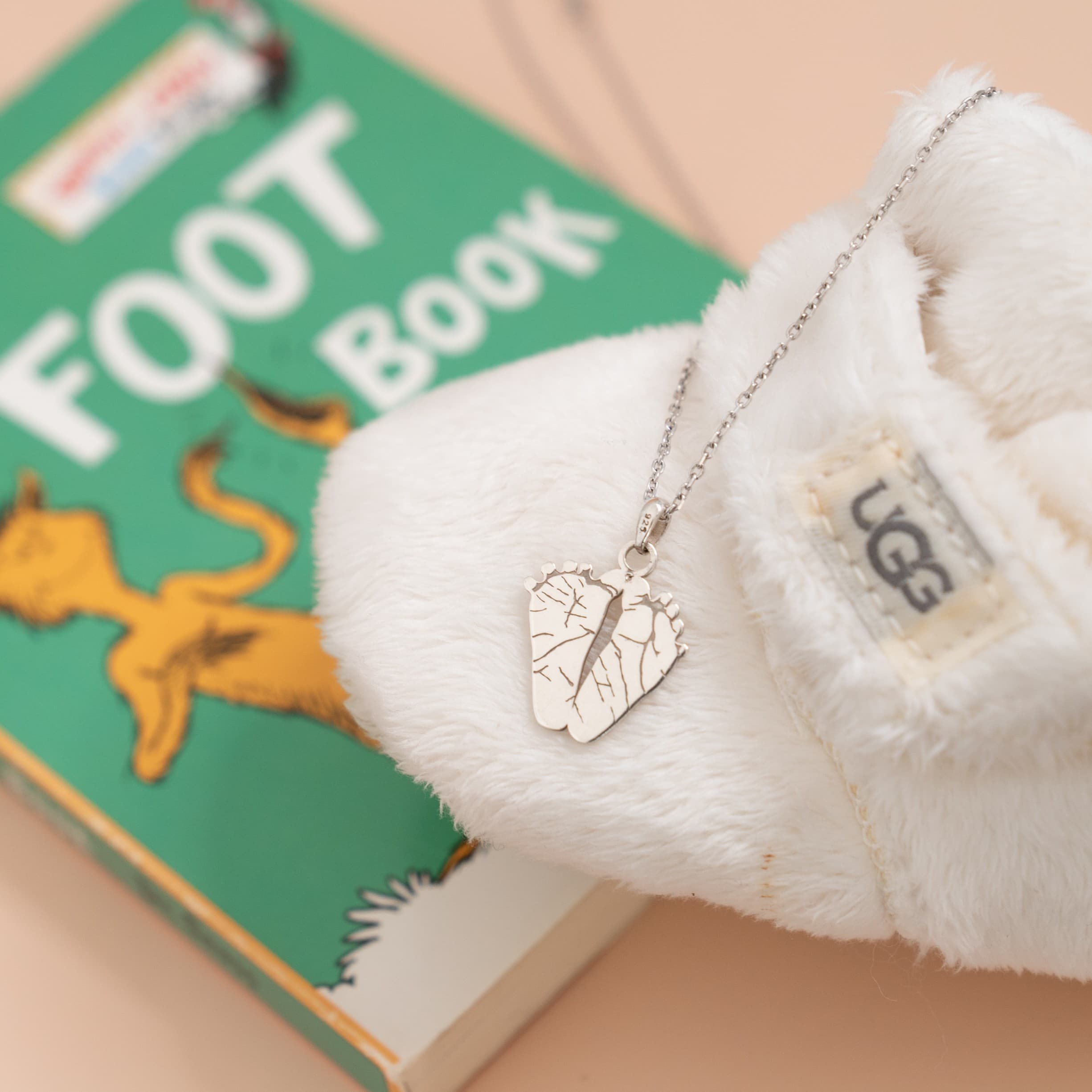 White UGG shoe with a silver necklace featuring an ilMio Footprint charm on top of a children's book